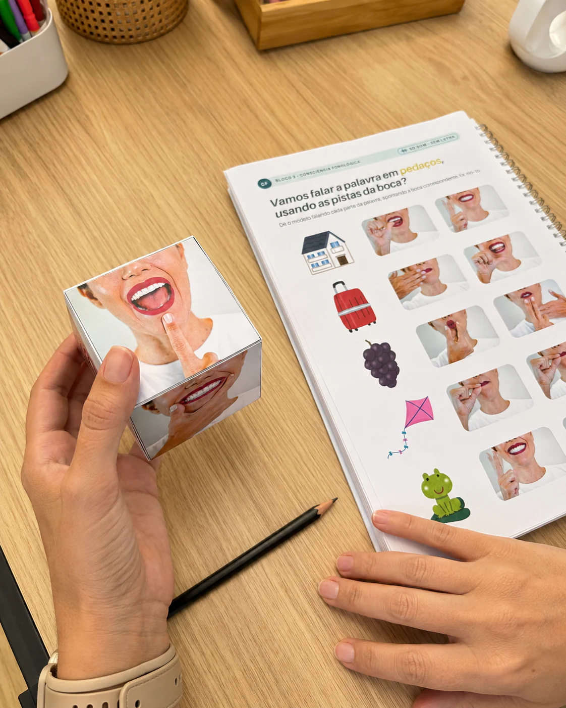 Mãos usando o dado das bocas montado e o caderno aberto numa mesa de atendimento