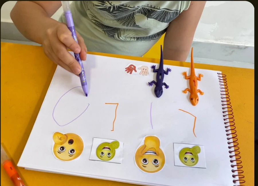Criança segurando a caneta e desenhando no caderno, com as bocas do MultiGestos Letícia coladas na página e brinquedos em cima da mesa